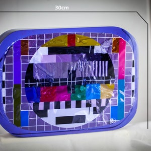 Op de afbeelding: Een rechthoekige paarse doos met een transparant plastic deksel. De doos heeft een zwart-wit televisie testpatroon erin. De doos is 30 cm breed en 22 cm hoog.