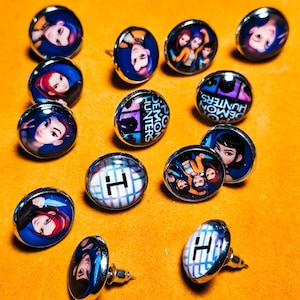 May include: A collection of silver-toned stud earrings with various designs. Some earrings feature images of characters, while others display the text "DEMON HUNTERS" or a white "H" on a blue and white checkered background. The earrings are arranged on an orange surface.