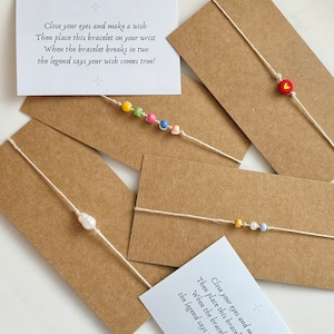 May include: Four friendship bracelets with different charms and beads. The bracelets are made of string and have a variety of colors, including red, pink, blue, yellow, green, and white. The charms include a heart, a pearl, and a small bead. The bracelets are displayed on brown paper cards with instructions on how to make a wish.