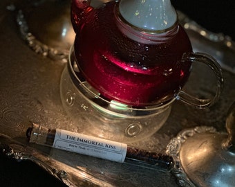 May include: A glass teapot filled with a deep red liquid sits on a silver tray. The teapot has a white and gold lid and is on a glass warmer. A small glass vial with the text "The Immortal Kiss" is next to the teapot.
