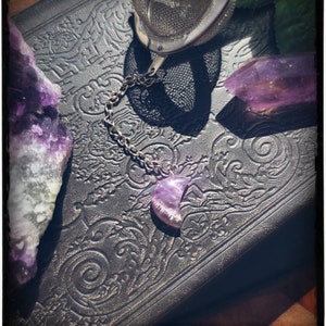 May include: A silver tea infuser with a chain and a purple amethyst crystal shaped like a crescent moon. The infuser is resting on a black leather-bound book with an ornate design.