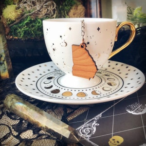 May include: A white teacup and saucer with a gold rim and a gold star pattern. A wooden tea infuser shaped like the state of Georgia hangs from the cup.