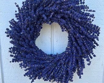 May include: A circular wreath made of dried lavender, showcasing a deep purple hue. The wreath is densely packed with lavender stems, creating a full, textured appearance. It hangs against a white wooden background.