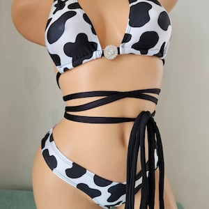 May include: A cow print bikini set. The halter-neck top has a central rhinestone detail. The bikini is white with black spots. Black straps wrap around the waist. The set is designed for swimwear.