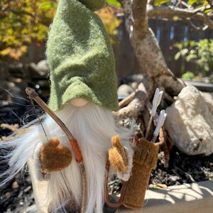 May include: A green felt gnome with a white beard and brown felt mittens holding a wooden bow and arrow. The gnome is wearing a brown felt quiver with arrows. The text "3. Archer" is visible below the gnome.