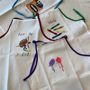 May include: Four white canvas aprons with colourful trim and embroidered designs. The aprons feature the phrases "love to colour", "World of", "love to paint", and "Art in progress".