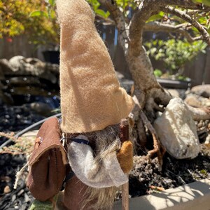 May include: A brown felt gnome with a tan hat and a brown backpack. The gnome is holding a wooden walking stick and is standing on a brown surface. The text "1. Hiker Side" is visible in the image.