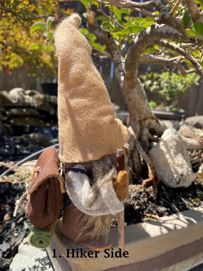 May include: A brown felt gnome with a tan hat and a brown backpack. The gnome is holding a wooden walking stick and is standing on a brown surface. The text "1. Hiker Side" is visible in the image.