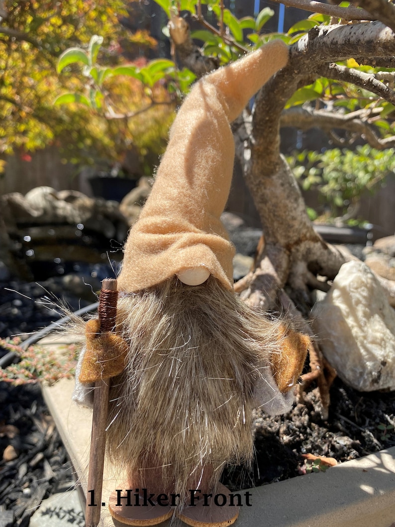 May include: A brown gnome with a long, shaggy beard and a brown felt hat. The gnome is holding a wooden walking stick and is standing on a brown surface. The text "1. Hiker Front" is visible in the image.