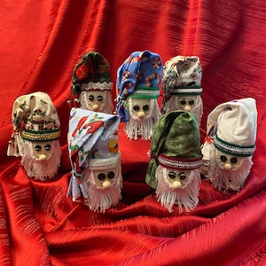 May include: Seven handmade Christmas gnomes with different patterned fabric hats and white beards. The gnomes are standing on a red fabric surface.