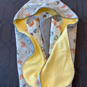 May include: A hooded towel with a yellow terrycloth lining and a grey cotton outer layer. The outer layer features a pattern of foxes, hedgehogs, and birds. The towel is folded in half and has a curved shape.