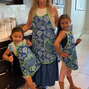 May include: Three people wearing blue and green floral print aprons are standing in a kitchen. The person in the middle is an adult woman, and the two people on either side are children. The children are holding kitchen utensils.