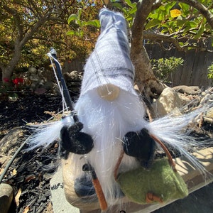 May include: A white and grey gnome with a long white beard, wearing a black hat and holding a fishing rod. The gnome is sitting on a wooden surface with a green bag.
