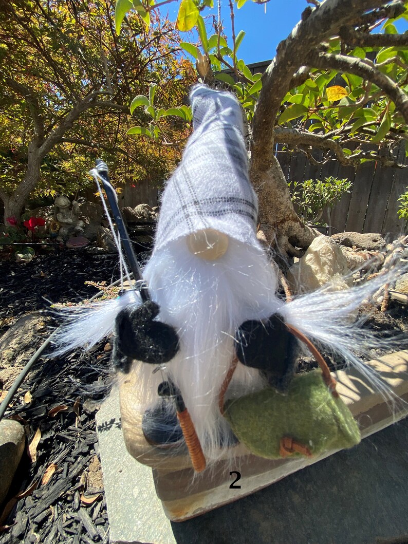 May include: A white and grey gnome with a long white beard, wearing a black hat and holding a fishing rod. The gnome is sitting on a wooden surface with a green bag.