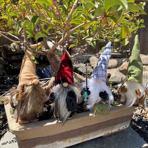 May include: Four gnomes with different coloured hats and beards are sitting in a brown planter. The gnomes are wearing different outfits, including a red and black plaid hat, a blue and white plaid hat, a brown hat, and a green hat. The gnomes are all holding fishing rods.
