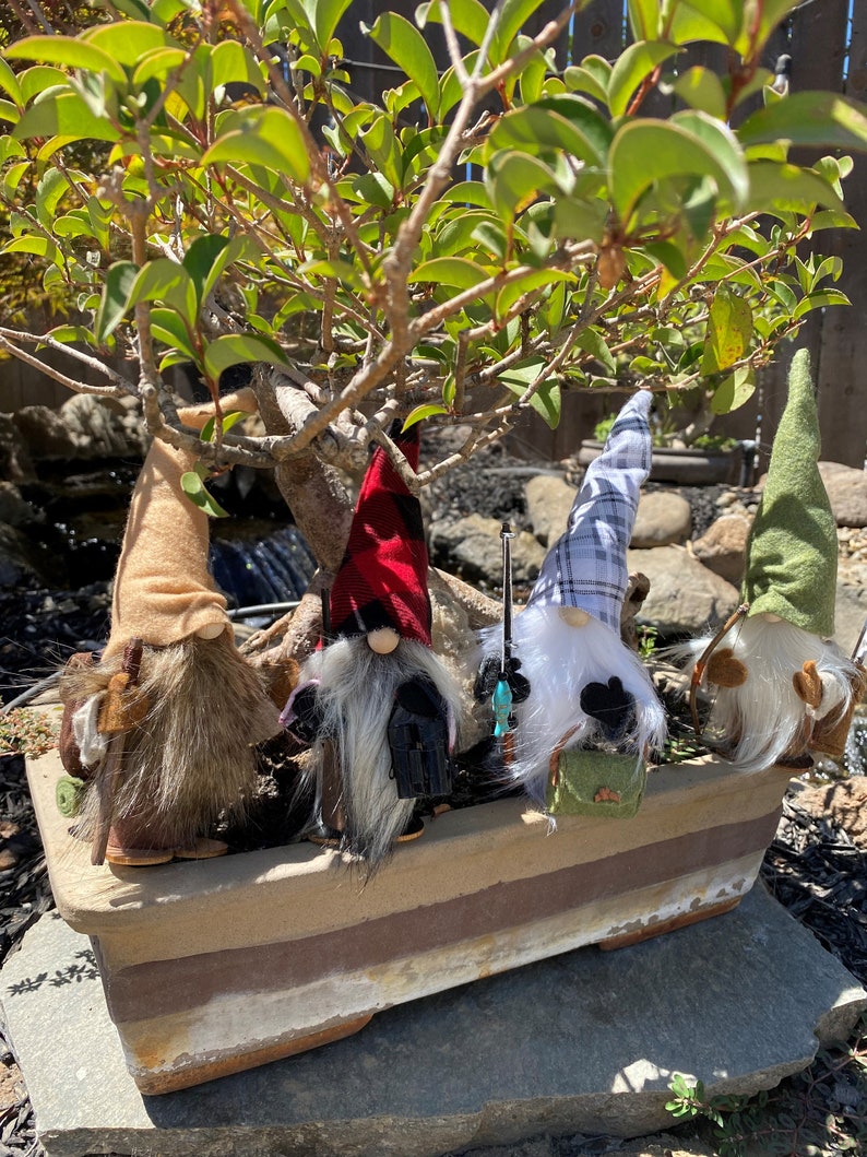 May include: Four gnomes with different coloured hats and beards are sitting in a brown planter. The gnomes are wearing different outfits, including a red and black plaid hat, a blue and white plaid hat, a brown hat, and a green hat. The gnomes are all holding fishing rods.