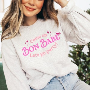 May include: A pale grey sweatshirt featuring the pink text "Come on Bon Babe Lets go party!" with a crown and starbursts. The sweatshirt is a relaxed fit, suitable for casual wear.