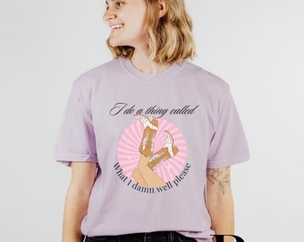 May include: A lilac-colored t-shirt featuring a graphic of cowboy boots and the text "I do a thing called what I damn well please." The design includes a pink circle with radiating lines. The shirt is a casual wear item.