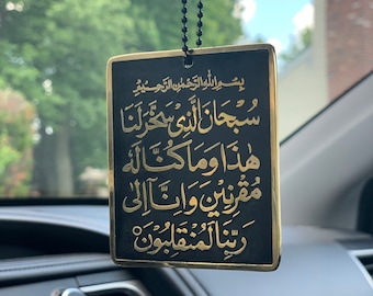 May include: A black and gold car air freshener with Arabic script. The rectangular pendant hangs from a black beaded chain. The text is in gold against a black background. The car's dashboard is visible in the foreground.