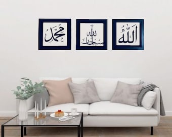 May include: Three framed wall art pieces with Arabic calligraphy in black on a white background. Each piece is framed in a dark blue square. The art is displayed above a white sofa with pillows.