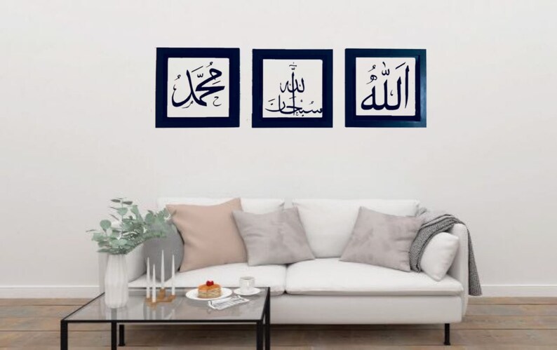 May include: Three framed wall art pieces with Arabic calligraphy in black on a white background. Each piece is framed in a dark blue square. The art is displayed above a white sofa with pillows.