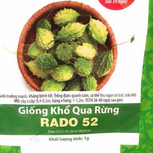 Könnte beinhalten: Eine Packung RADO 52 Bittermelonen-Samen. Die Packung zeigt ein Nahaufnahmebild von frischen Bittermelonenfrüchten in einem geflochtenen Korb. Der Text auf der Packung lautet "Giống Khổ Qua Rừng RADO 52" und "Khối lượng tịnh: 1g".