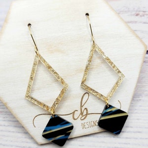 May include: Gold geometric dangle earrings with black and blue striped resin accents. The earrings feature a gold diamond shape with a smaller diamond shape hanging from the bottom.