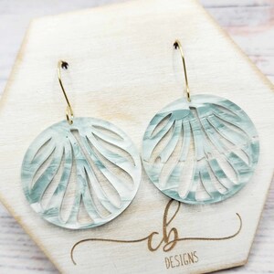 May include: A pair of light blue, round, openwork earrings with a marbled effect. The earrings are hanging from gold hooks.