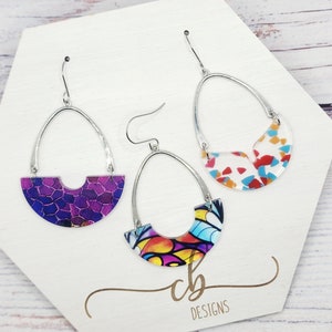 May include: Three pairs of dangle earrings with silver hoops and colorful, abstract, resin-like designs. The earrings are displayed on a white hexagonal stand with the text "cb Designs".