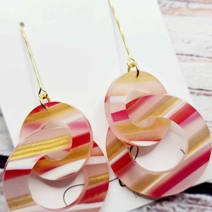 May include: A pair of dangle earrings with gold hooks and two overlapping circles. The circles are made of translucent pink, white, and gold striped acrylic.
