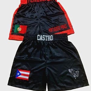 Puede incluir: Dos pares de pantalones cortos de boxeo de satén negro. Un par tiene "FERREIRA" en rojo en la cintura y el texto "PORTUGUESE PRINCE" en el lateral, con la bandera portuguesa. El otro par tiene "CASTRO" en la cintura y la bandera puertorriqueña.