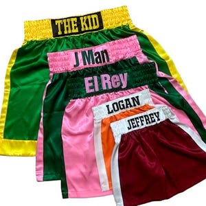 Könnte beinhalten: Eine Ansammlung von Boxshorts aus Satin in verschiedenen Farben, darunter Grün, Rosa, Orange und Burgunderrot. Jede Hose hat einen Namen auf dem Bund gestickt: "THE KID", "J Man", "El Rey", "Logan" und "Jeffrey".