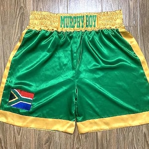 Op de afbeelding: Groene boksshorts met gouden randen en de tekst "MURPHY BOY" over de tailleband. De shorts hebben een Zuid-Afrikaanse vlag patch op het linkerbeen. De shorts zijn gemaakt van een glanzend, satijnachtig materiaal.