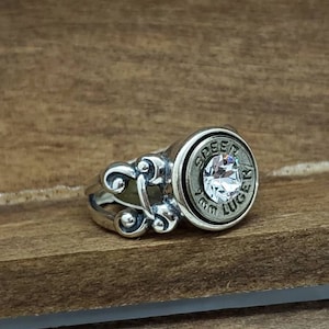 May include: A silver ring with a bullet casing design. The bullet casing has a clear crystal in the center and the words "9mm Luger" engraved around the edge.
