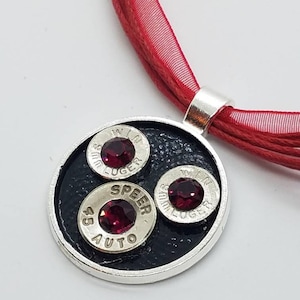 May include: A silver pendant necklace with a black circular background and three bullet casings with red gemstones. The necklace is on a red cord.