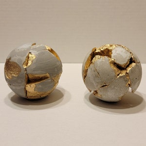 May include: Two gray concrete spheres with gold accents. The spheres are cracked and broken, revealing the gold underneath.