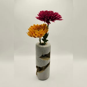 May include: A grey concrete vase with a distinctive cracked pattern displays two flowers. One is a bright orange, the other a deep burgundy. The cylindrical vase has a rough, textured surface, adding a modern, artistic touch to the floral arrangement.