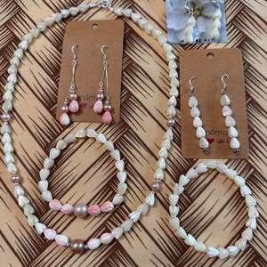 Könnte beinhalten: Ein Set aus weißem Muschel-Schmuck mit rosa Blumenakzenten. Das Set beinhaltet eine Halskette, ein Armband und ein Paar Ohrringe. Der Schmuck ist handgefertigt mit Liebe.