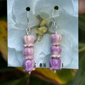 May include: A pair of dangle earrings featuring a floral design. Each earring has three stacked, bell-shaped beads in shades of pink and purple, suspended from a silver-colored hook. The earrings are displayed on a floral-themed card.