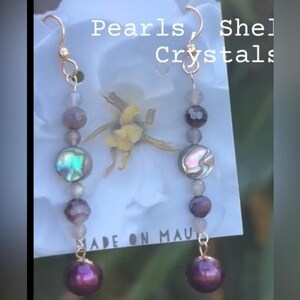 May include: Gold-coloured earrings with pearls, shell, and crystals. Each earring has a gold hook, small beads, an iridescent shell disc, and a large, deep purple pearl. The text "Pearls, Shell, Crystals" is visible.