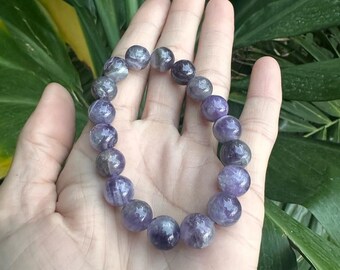 May include: A bracelet featuring polished amethyst beads, predominantly purple with greyish translucent sections. The beads are strung together in a circular form. The bracelet is shown on an open hand, set against a background of green leaves.