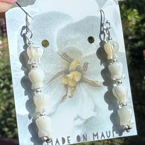 May include: A pair of white shell earrings with silver accents. The earrings are hanging from a white card with the text "MADE ON MAUI".