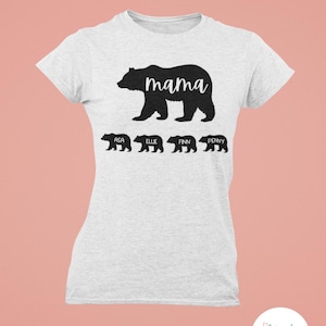 May include: A light gray t-shirt featuring a black bear graphic with the word "mama" and four smaller bear silhouettes below, each with a name: Asa, Ellie, Finn, and Penny. The shirt has a crew neck and short sleeves.