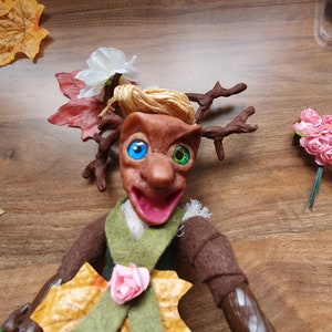May include: A handmade doll with brown skin, green and blue eyes, and a wide smile. The doll is wearing a green and brown outfit with a pink flower and a large brown leaf. The doll has antlers made of brown branches and blonde hair.