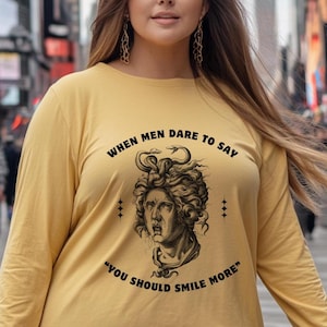May include: A yellow long-sleeved shirt with a black graphic of a woman's face with snakes in her hair. The text on the shirt reads "When men dare to say "You should smile more."