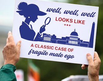 May include: A white sign with blue and red text reads "well, well, well LOOKS LIKE A CLASSIC CASE OF fragile male ego". A blue silhouette of a woman with a magnifying glass is in the upper left corner.