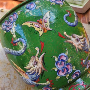 Peut inclure: Un bol décoratif en métal vert avec un design détaillé. Le bol présente un motif de dragon et de papillon avec des accents floraux roses, bleus et jaunes. Le bol a une bordure dorée et une finition brillante.