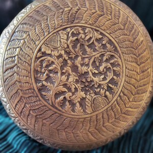Pode incluir: Close-up detalhado de um objeto redondo de latão antigo. A superfície é intrincadamente esculpida com padrões florais e de folhas, criando um design texturizado e ornamentado. A cor do objeto é um dourado quente e envelhecido.