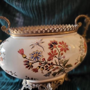 Peut inclure: Un bol décoratif en céramique blanche avec des motifs floraux et de libellules. Le bol a des poignées dorées et un bord décoratif. La base est également dorée. Le bol mesure environ 25 cm de large.