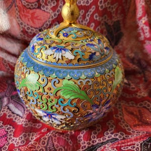 May include: A round, ornate cloisonné jar with a lid. The jar features a gold base with intricate floral designs in blue, green, white, and red enamel. The lid is topped with a gold finial. The background is a red patterned fabric.
